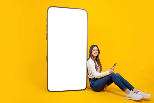 Full length body view woman sitting leaning big huge blank empty white screen mobile phone mockup. Caucasian teen girl holding smartphone. Smiling, recommending application, isolated yellow background