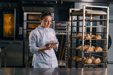 a woman baker in a uniform near the shelves with freshly baked bread holds a tablet in her hands checks bread bakery