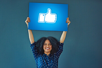 Portrait, social media and thumbs up icon to like with a woman holding a poster in studio on a blue...