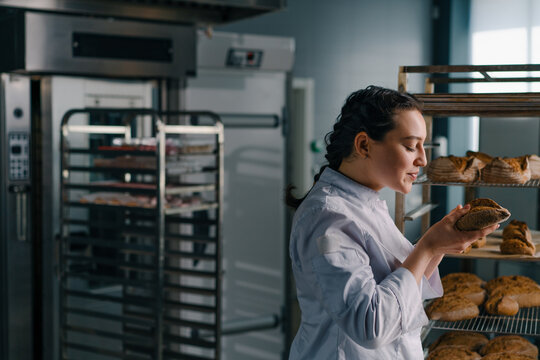 attractive female baker takes freshly baked fragrant bread from the shelf sniffs it checks its quality bakery professional kitchen baking