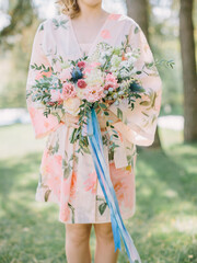 A bride in a pink robe in the garden holds a wedding bouquet with echeveria, pink and white peonies and roses. Blue and pink ribbons hang from the bouquet and develop. Wedding day.