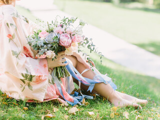 A bride in a pink robe with flowers sits on the lawn and holds a wedding bouquet with echeveria, pink and white peonies and roses. Bouquet tied with ribbons.