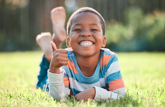 Thumbs up, portrait of black boy and lying on grass in a nature park with a lens flare. Good news or thank you, success or winner and happy or excited young male child relaxing on a lawn outdoors