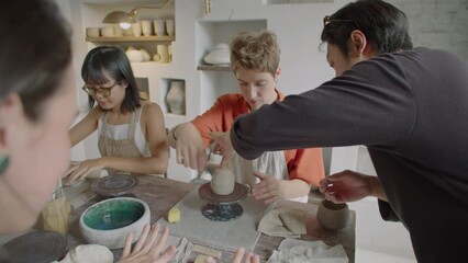 Professional ceramist teaching girl how to work with pottery wheel while giving pottery masterclass to group of female students in workshop