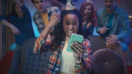 An African American girl is playing an online game on her phone while her friends are watching her from behind her. The girl wins, waving her hand in a victory gesture. Close up.