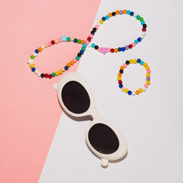 Round white plastic sunglasses, necklace and bracelet made of colored beads on a pink and white background.