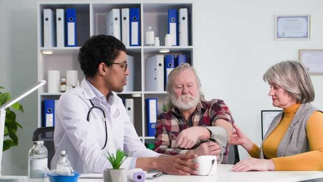 young afro american doctor measures pressure of an elderly man with tonometer, an old woman rejoices in good health of her husband