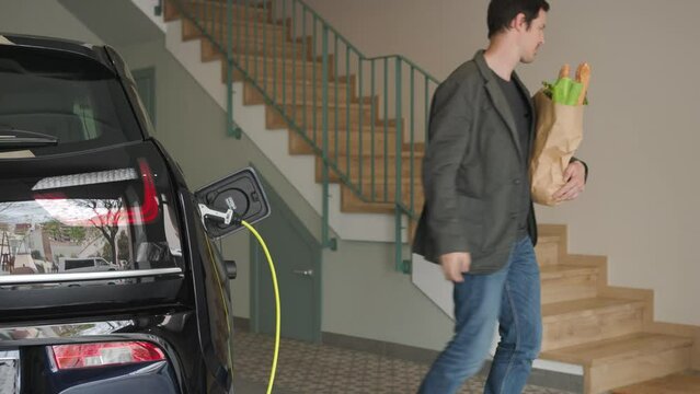 From behind, the man carrying the shopping bag connects a charger to the socket of an electric blue car and goes up the stairs to his house. sustainability efficiency Home Living. 4k horizontal video