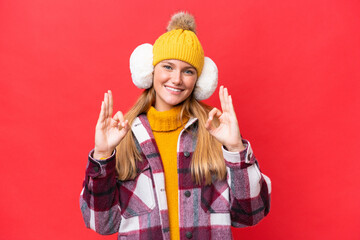 Young beautiful woman wearing winter muffs isolated on red background showing an ok sign with fingers