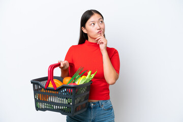Plakat Young Asian woman holding a shopping basket full of food isolated on white background having doubts while looking up