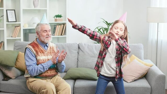 Playful young lady with birthday cone on head dancing like rock star in bright living room. Cheerful granddad enjoying performance, clapping hands and showing thumb up to beloved grandchild.