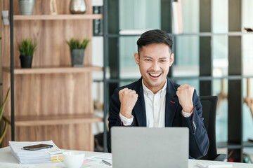 Successful and happy young Asian man, businessman, happy, shows with hand yes. Made a contract. Won money. Sitting with a smart phone or notebook at the table in a modern office.