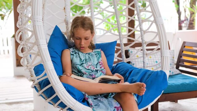 Little girl reading book while resting in hanging swing chair on tropical terrace