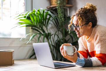 Happy adult woman using laptop at home and drinking coffee. Modern lady smiling at the computer screen in video chat connection leisure online activity. People at home surfing the net and enjoying day