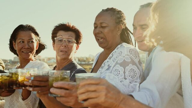 Happy multiracial senior friends having fun drinking and toasting mojitos on the beach during sunset time - Elderly people enjoying summer holidays