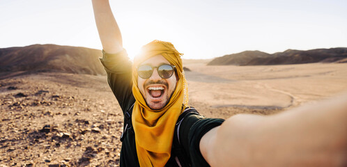Millennial man taking selfie pic with smart mobile phone in rocky desert - Traveler guy with backpack enjoying day out on summer vacation - Happy tourist having fun at summertime holiday - obrazy, fototapety, plakaty