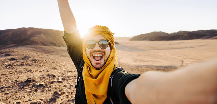 Millennial man taking selfie pic with smart mobile phone in rocky desert - Traveler guy with backpack enjoying day out on summer vacation - Happy tourist having fun at summertime holiday