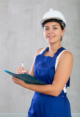 Portrait of emotional mechanic girl in blue overalls and a protective helmet, who writes in documents