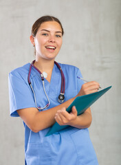 Positive polite young woman general practitioner in blue uniform filling out clipboard with medical records. Studio photoshoot on gray background - obrazy, fototapety, plakaty