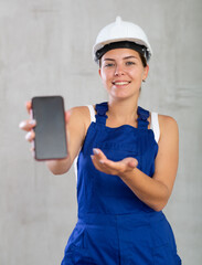 Closeup of professional smiling female engineer, construction manager in blue construction overalls and safety helmet, showing smartphone screen, grey background