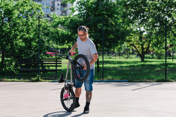 A cool middle-aged tattooed man is standing in a skate park with his bmx bike and preparing to perform freestyle tricks.