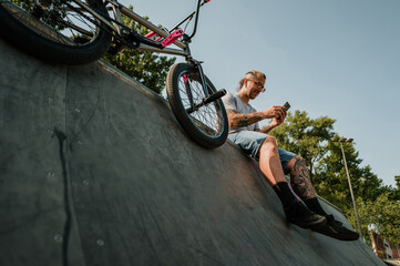 Low angle view of an urban tattooed mature man sitting in a skate park and scrolling on the phone.