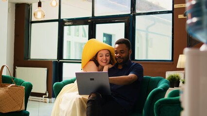 Couple planning trip itinerary on laptop while they wait to check in at hotel, lounge area. Diverse tourists browsing website and social media app to find leisure activities at seaside.