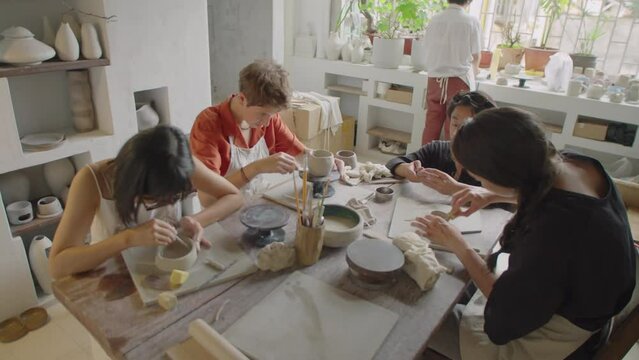 High angle shot of group of female students sitting around table and working with clay with assistance of Asian male teacher during pottery workshop