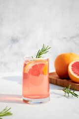 Elegant glass with a fresh, cooling cocktail of fresh grapefruit or red orange with ice cubes and a sprig of rosemary on a gray background on a bright sunny day. Vacation and summer drinks concept.