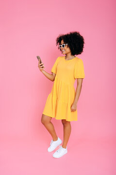 Full-length of happy latino or brazilian woman, in yellow sundress, using her mobile phone, chatting online with friends, browsing internet, texting, looks at a screen, smile, isolated pink background