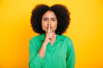 Fototapeta na wymiar Secret, mystery. Photo of pretty brazilian or latino woman, wearing casual wear, showing shh sign with forefinger touching her lips, stop talking, looks at camera, stand on isolated yellow background