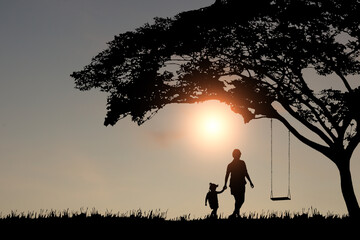 A silhouette of a mother holding her daughter's hand to walk to the swing under a big tree in the...