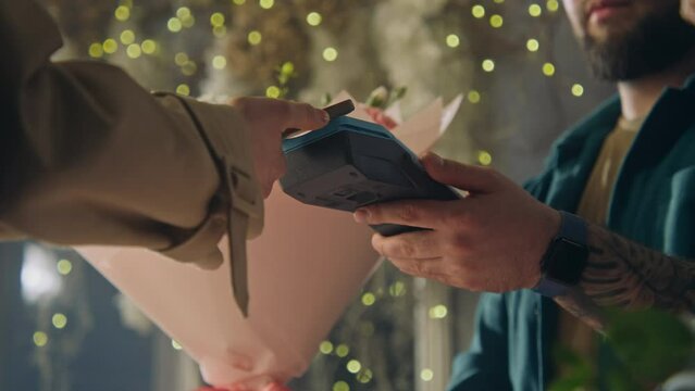 Male florist, seller gives payment terminal to female buyer. Woman pays for flowers bouquet in wrapping paper with contactless payment using phone in flower shop. Floristry, small business. Close up.