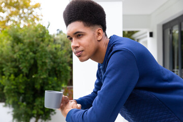 Thoughtful biracial man standing on balcony, looking away and holding mug, unaltered