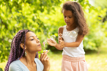 Obraz premium mother with daughter in the park, african young woman with child playing with soap bubbles in nature, family spending time together, happy kid with single parent