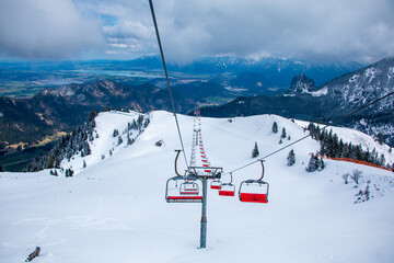 Fototapeta na wymiar Empty red chairlift near the summit of Mount Breitenberg in the German Alps, surrounded by contrasting wintry landscape up high and green meadows in the valleys below