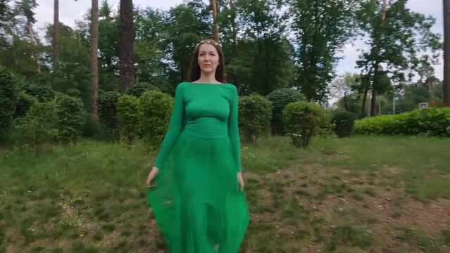 A fairy woman in a green dress walks on the grass in the park.