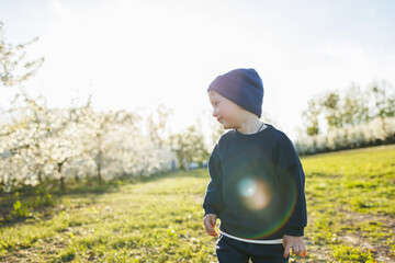 A little boy, 3 years old, in a sweater and a hat, runs through a blooming garden. Clothes for...