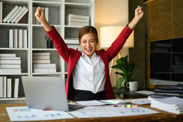 Excited business woman in red suit looking at laptop screen, making winner yes gesture, celebrating business success