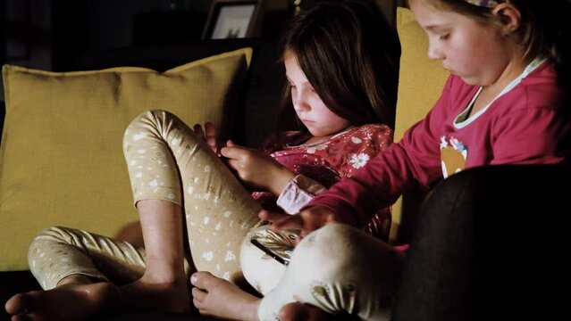 Side view of twin girls engrossed with digital devices on couch, new age kids