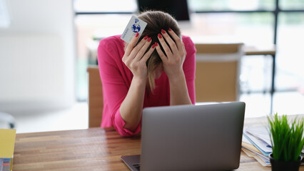 Upset woman sitting at laptop with credit plastic card and holding his head. Internet scam concept