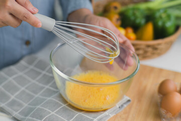 Young woman cooking in bright kitchen, hands whisking eggs in a bowl placed on towel and wooden table. Preparing ingredients for healthy cooking. Homemade food