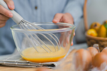 Young woman cooking in bright kitchen, hands whisking eggs in a bowl placed on towel and wooden table. Preparing ingredients for healthy cooking. Homemade food
