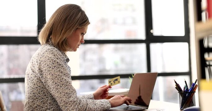 Focused woman enters credit card details on shop website via laptop sitting at table in office. Manager makes online payment at work slow motion