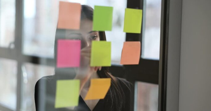 Female secretary writes on stickers important work plans on glass board by large window in office premise. Woman employee fills stickers to remind of events slow motion