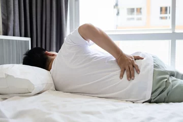 Fotobehang Stressed asian man having a backache,sore hips,waist hurts,unhappy adult people suffering from low lumbar pain or acute back strain,lying on the mattress at home,health care,medical,lifestyle concept © Satjawat
