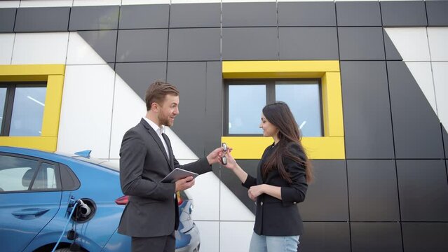 Man dealer with tablet passing car keys to happy owner outdoors. Cheerful beautiful Caucasian woman turning to camera and bragging purchase of automobile outdoors