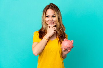 Teenager blonde girl holding a piggybank over isolated blue background looking to the side and smiling