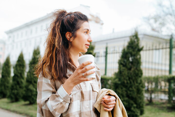 Portrait woman coffee break in park. Young woman holding raincoat and disposable coffee cup while walking outdoors and looking away