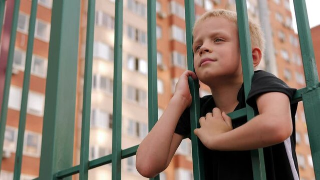 Close-up of a little boy standing behind a green metal fence on a sports field. A child walks and plays on the playground against the background of a multi-storey building.
